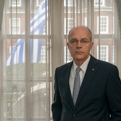 Ambassador Dimitris Caramitsos-Tziras standing in front of the Greek flag